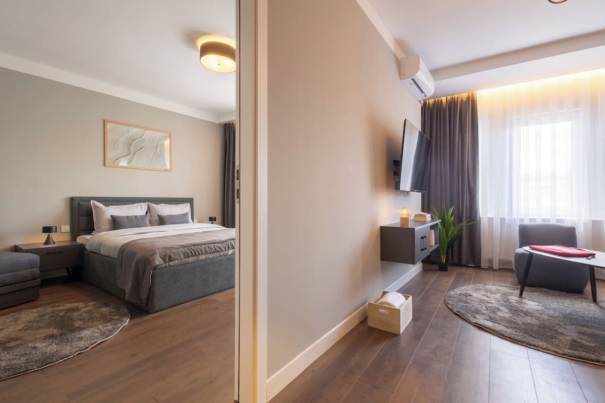 A modern hotel room with a bed, bedside table, TV, and a seating area with a table and chairs.