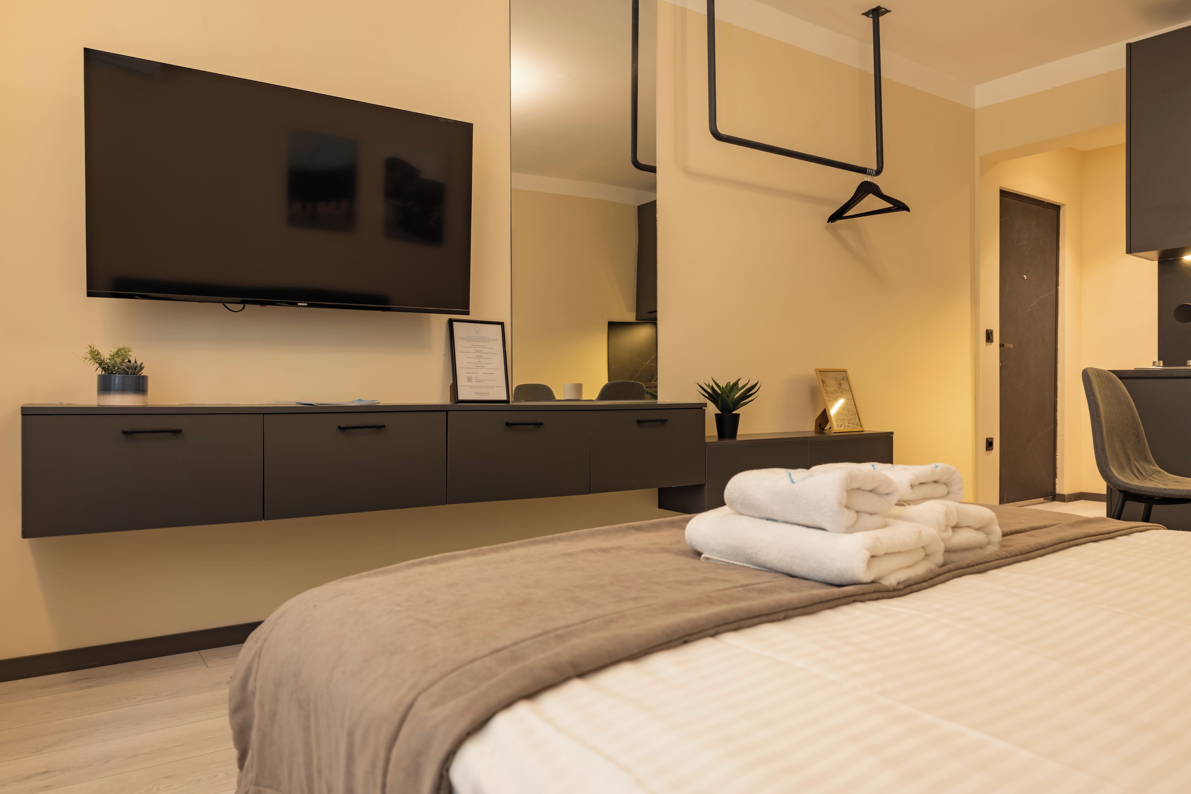 A modern bedroom with a flat-screen TV mounted on the wall, a large bed with neatly folded towels, a chair, and a desk with a plant.