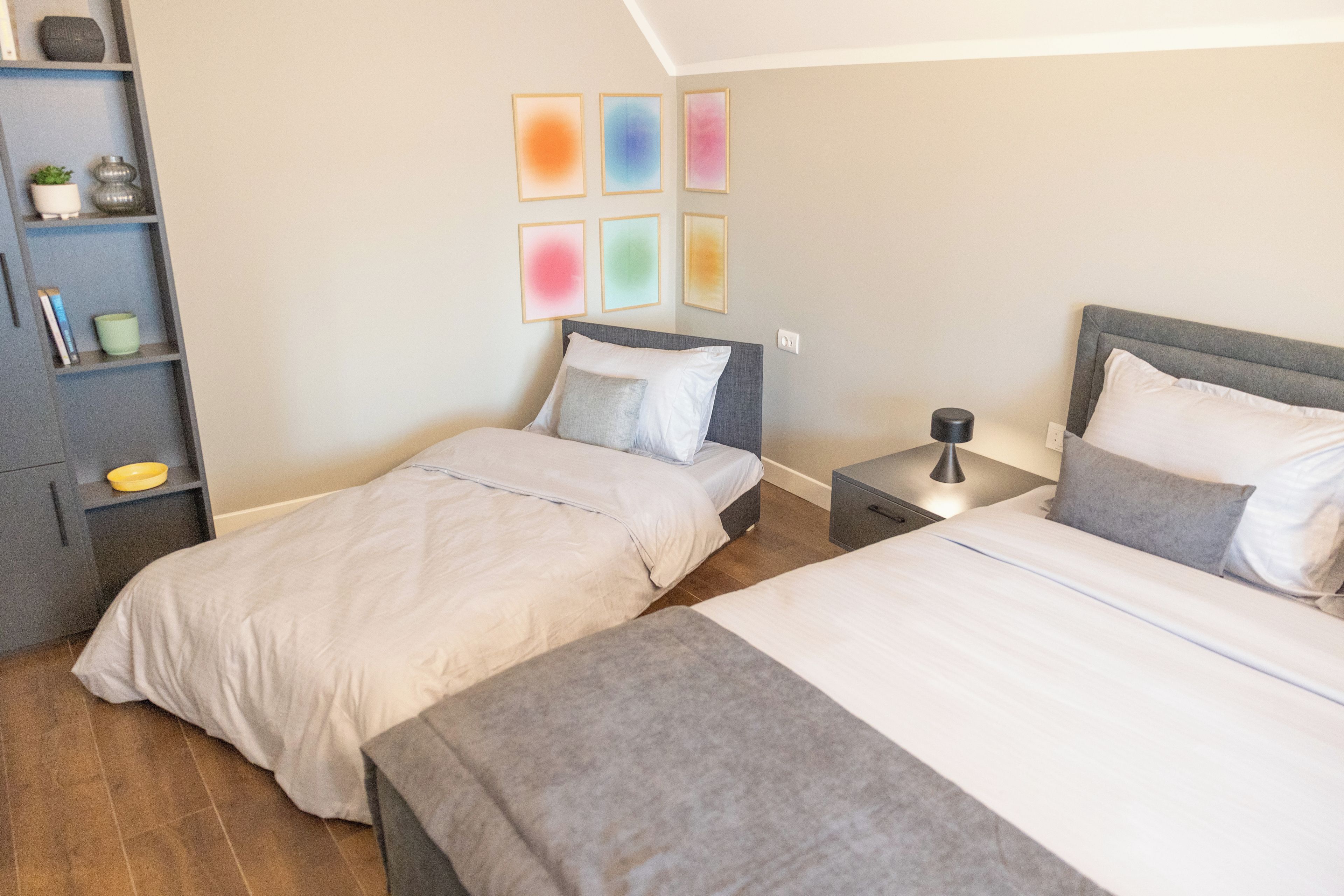 Ein Schlafzimmer mit zwei Betten, einem Nachttisch, einem Regal mit Dekoration und einer Wand mit farbenfroher Kunst.