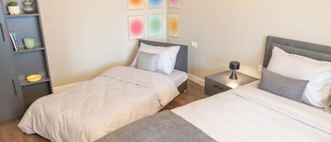 A bedroom with two beds, a bedside table, a nightstand, a shelf with decorative items, and a wall with colorful artwork.