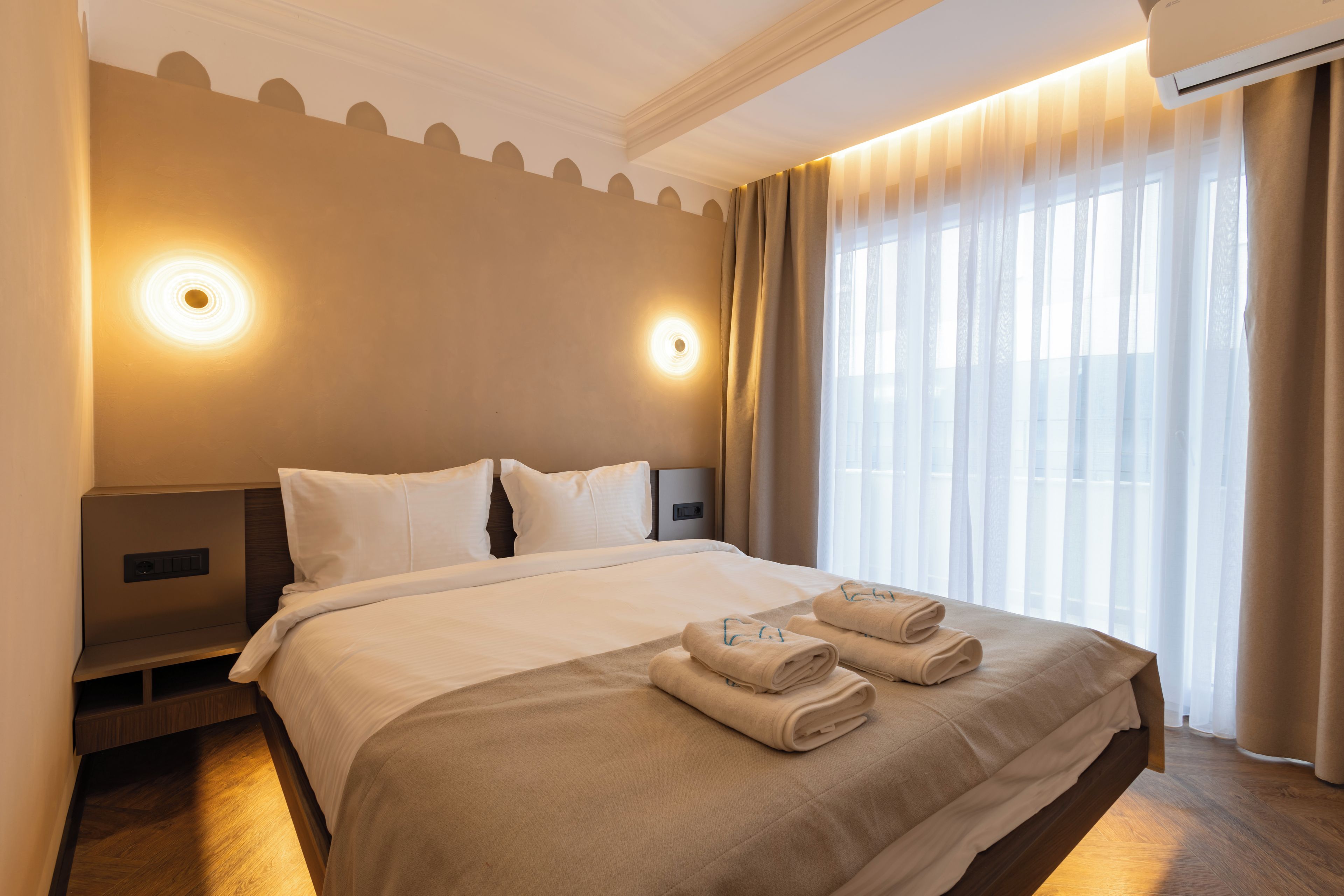 A hotel room with a large bed, two bedside tables, a nightstand, a window with sheer curtains, and wall-mounted lights.