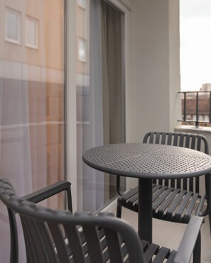 A balcony with a round table and two chairs, a window with blinds, and a view of buildings. - Prishtina City Apartments (Pristina)