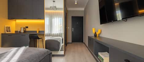 A modern hotel room with a bed, a desk, a TV, and a kitchenette.