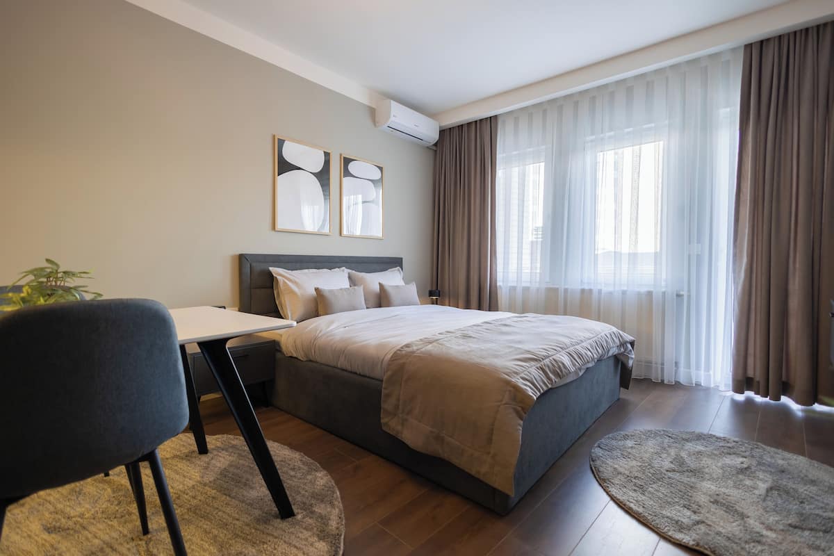 A modern hotel room with a large bed, a desk, and a chair. There is a window with curtains, a plant, and artwork on the wall.