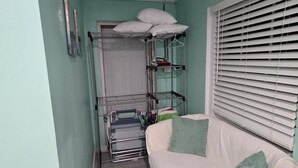 A narrow room with a white sofa, a metal shelf unit, and a window with blinds. - Centrally located 1 bedroom efficiency (georgetown)
