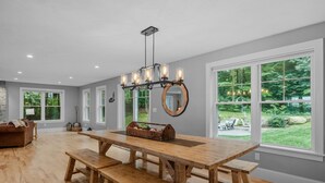 A spacious kitchen with a large wooden table and benches, a chandelier, and large windows offering a view of the outdoors. - Sunset Retreat at Dockham Shore (Gilford)