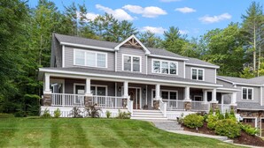 A two-story house with a front porch, a covered entryway, and a landscaped front yard. - Sunset Retreat at Dockham Shore (Gilford)