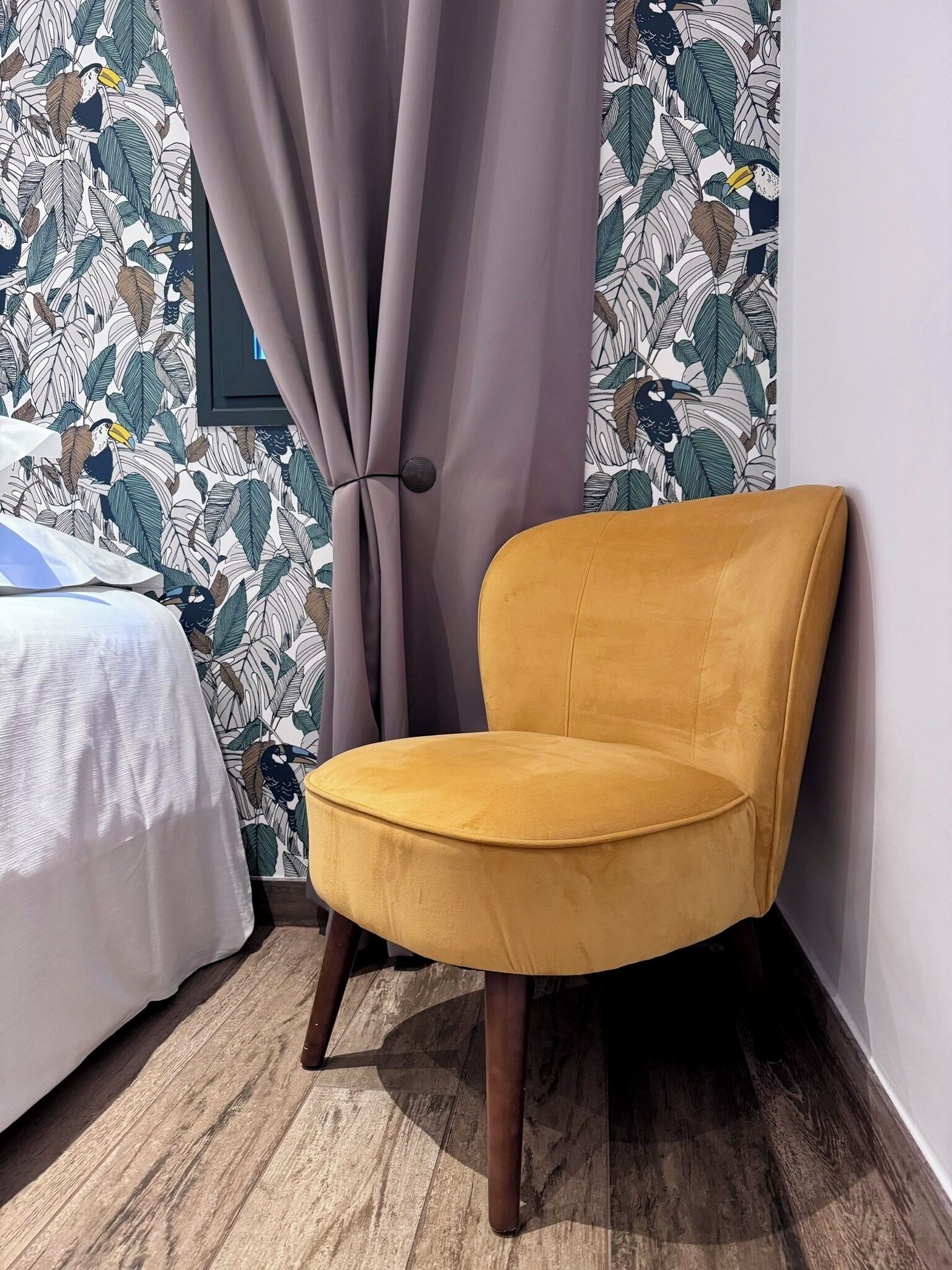 Ein Teil eines Zimmers mit einem gelben Sessel, einem Bett mit weißer Bettwäsche und einer Wand mit einem Blättermotiv.