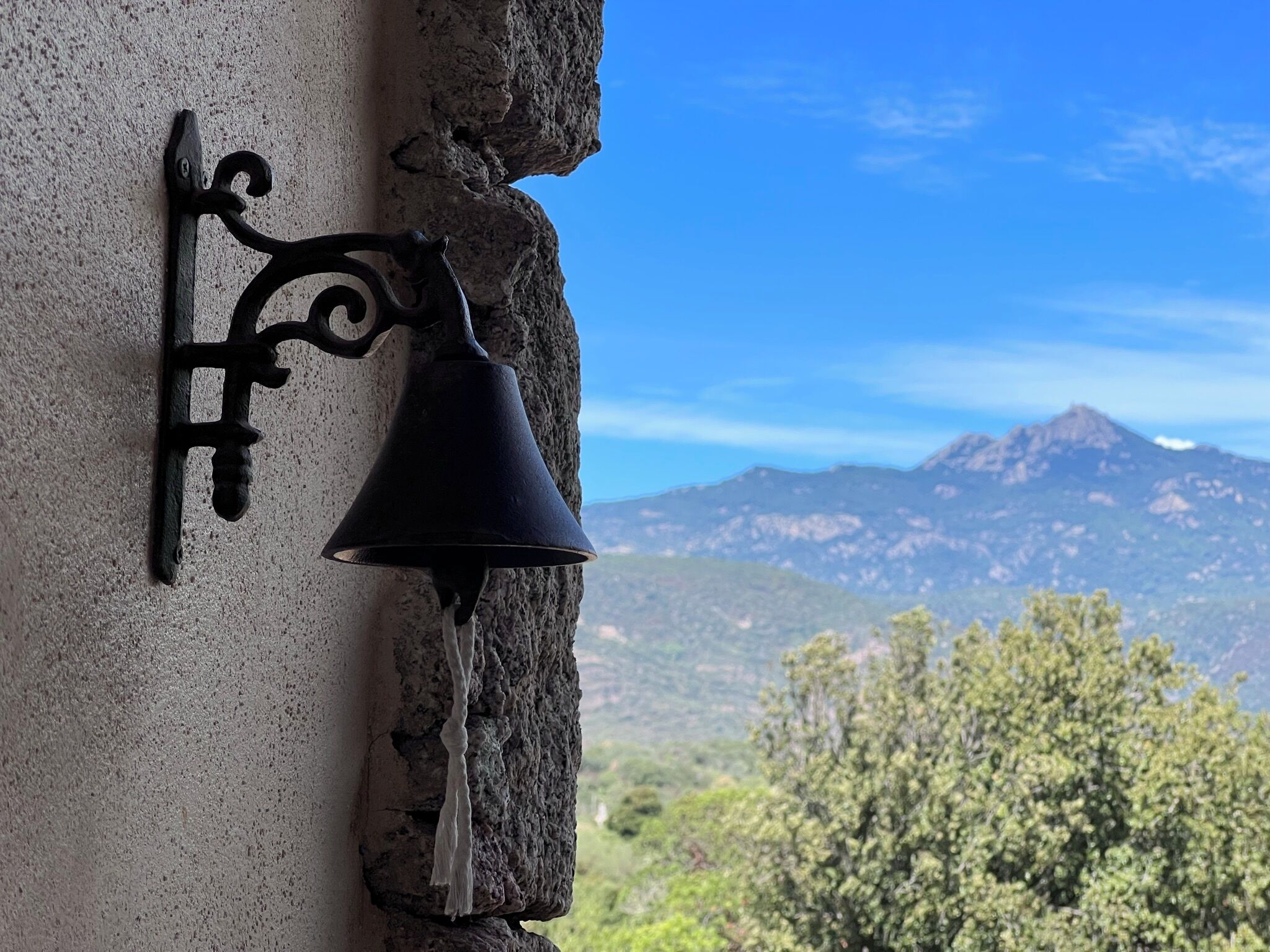 Un mur de pierre surmonté d’une cloche en métal noir, avec une vue sur les montagnes en arrière-plan.