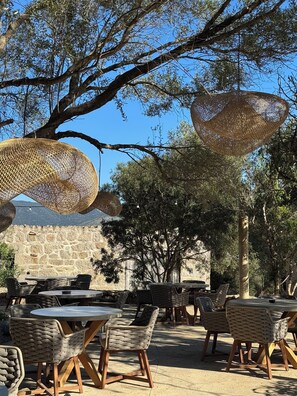 An outdoor seating area with wicker furniture and hanging rattan lamps, set against a backdrop of trees and a stone wall. - Hôtel confidentiel Le Grécale **** (Pianotolli-Caldarello)