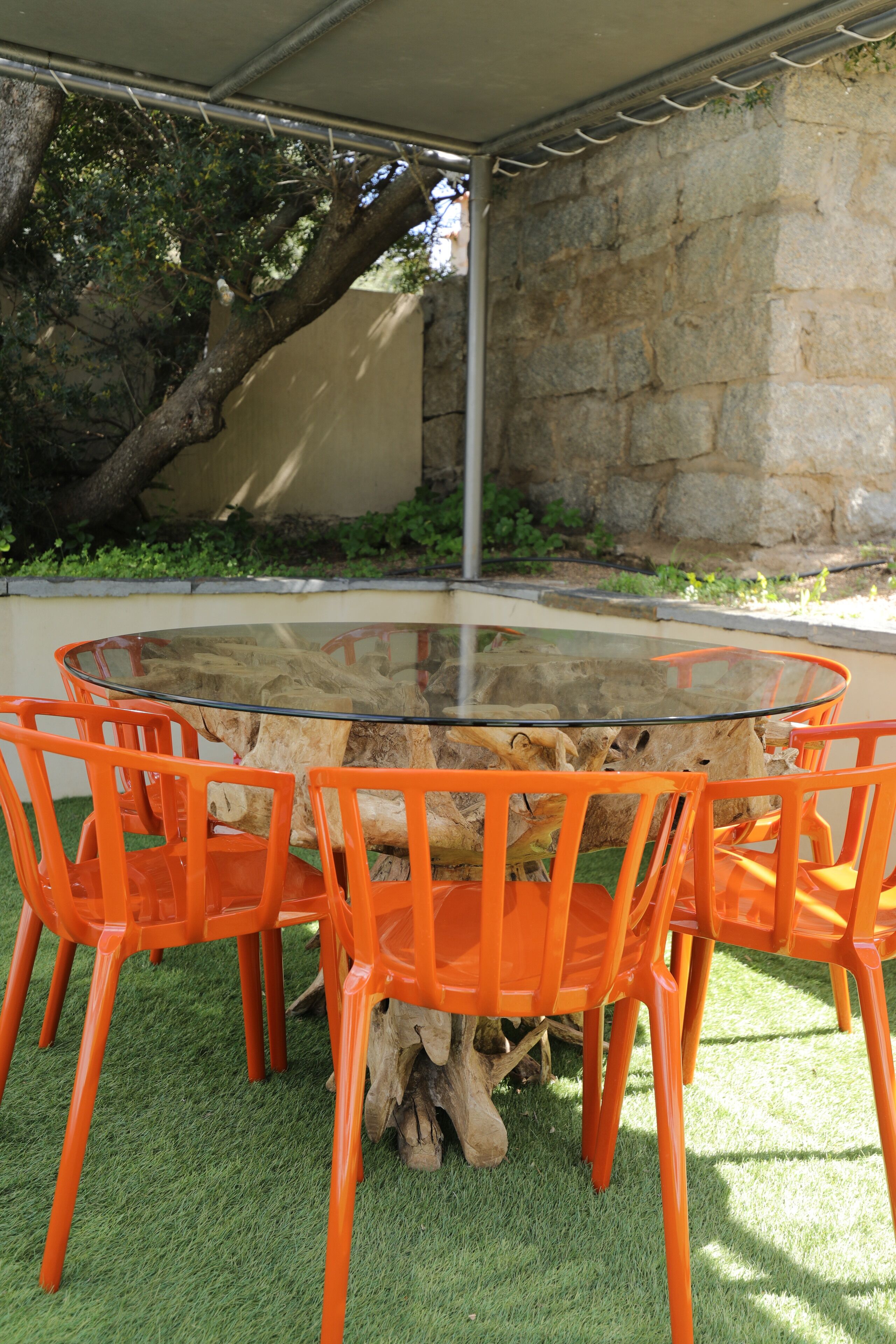 Una mesa de vidrio redonda rodeada por seis sillas naranjas, sobre césped artificial, con un muro de piedra y un árbol al fondo.
