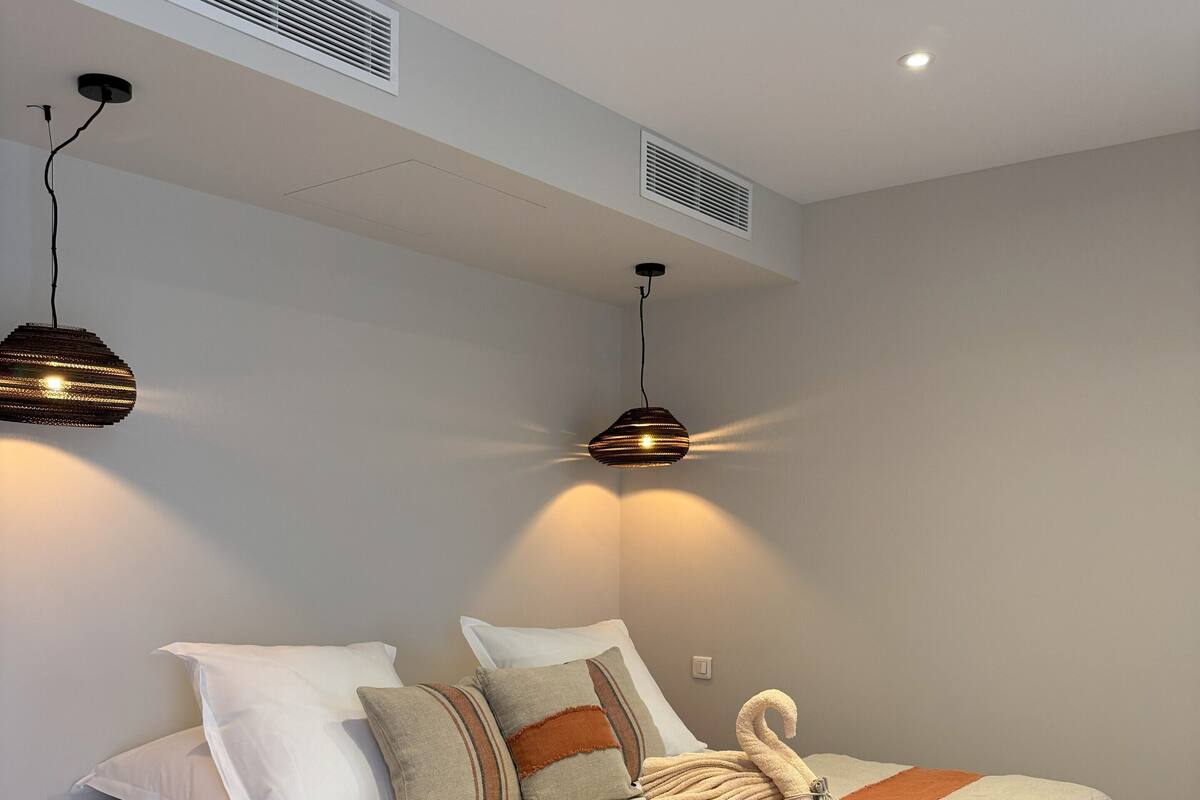 Egy modern hálószoba, melyben egy nagy ágy, két éjjézőlámpa, egy mennyezeti ventilátor és egy beépített szellőzőnyílás található.