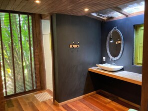 A bathroom with a wooden vanity, a large mirror, and a view of greenery through a window with blinds.