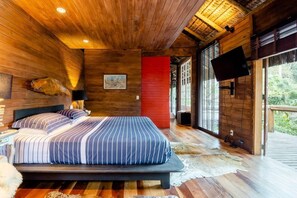 A wooden cabin bedroom with a bed, a red accent wall, a mounted TV, and large windows. - Rainforest Paradise (Rio de Janeiro)