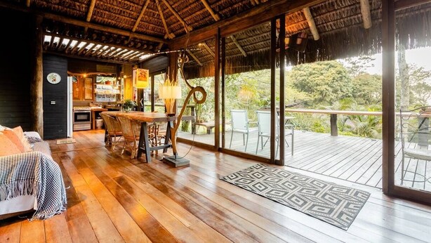 Una habitación amplia con piso de madera, techo de paja y un gran ventanal corredizo que da a una terraza exterior.
