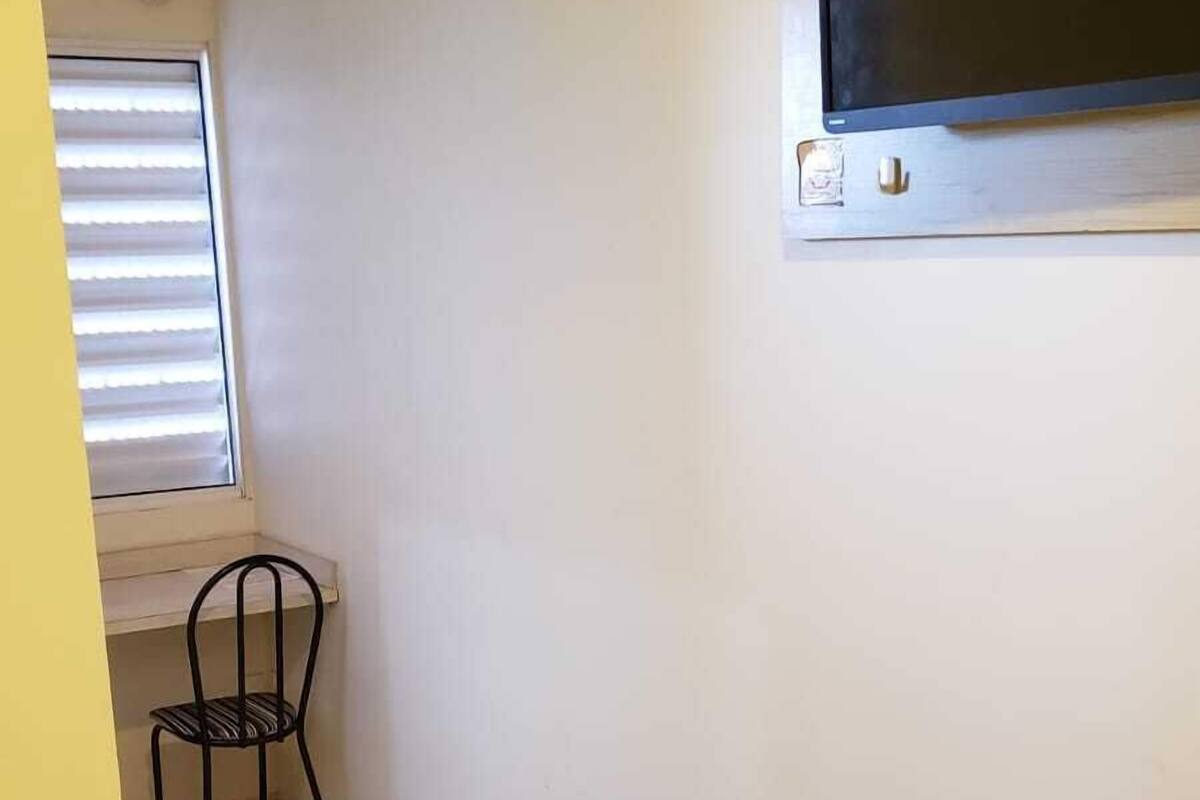 Une petite chambre d’hôtel avec deux lits, un téléviseur fixé au mur, une fenêtre avec des stores et une chaise.