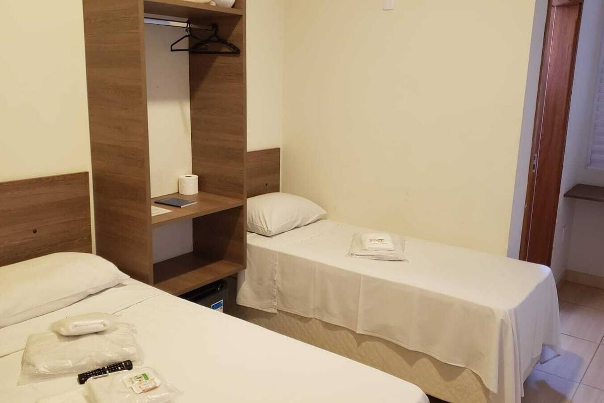 Une petite chambre d’hôtel avec deux lits, une tête de lit en bois, une étagère avec un manteau suspendu et une fenêtre avec des stores.