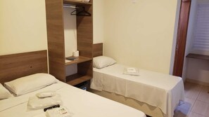 A small hotel room with two beds, a wooden headboard, a shelf with a hanging coat, and a window with blinds. - Hotel Viracopos de Indaiatuba Ltda EPP (Indaiatuba)