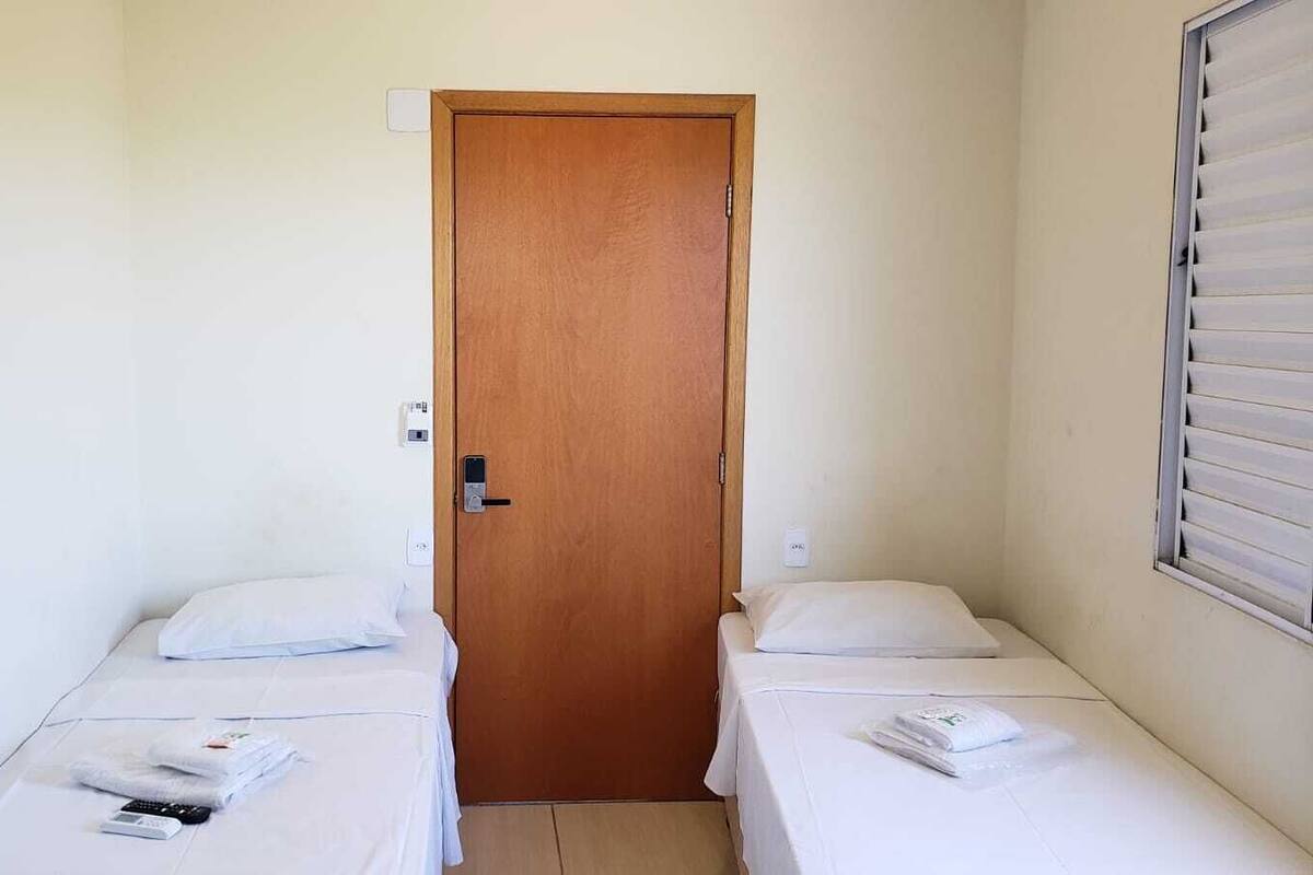 Une petite chambre propre, avec deux lits simples, une porte en bois et une fenêtre avec des stores.