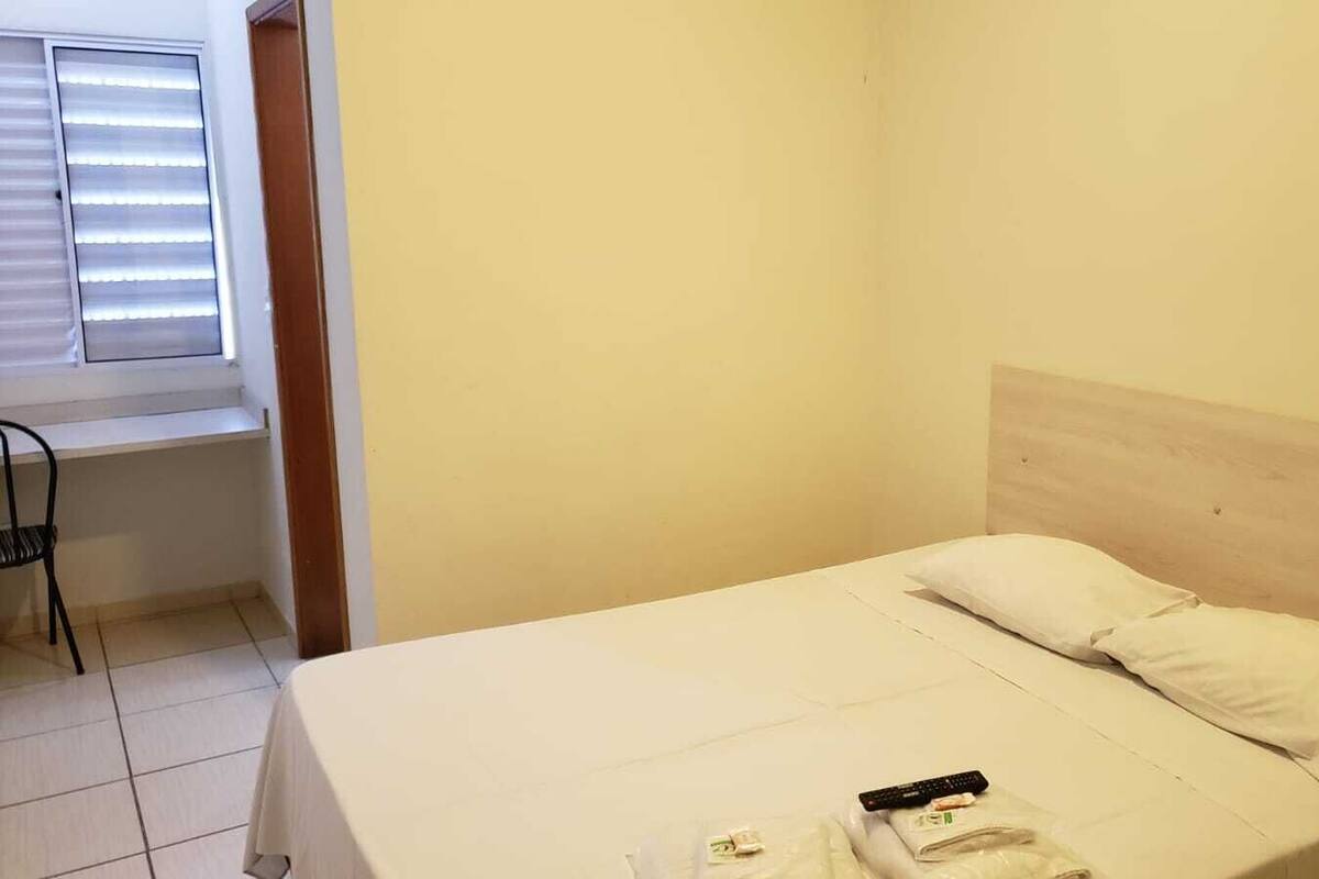 Une petite pièce propre, avec un lit simple, une chaise et une fenêtre avec des stores.