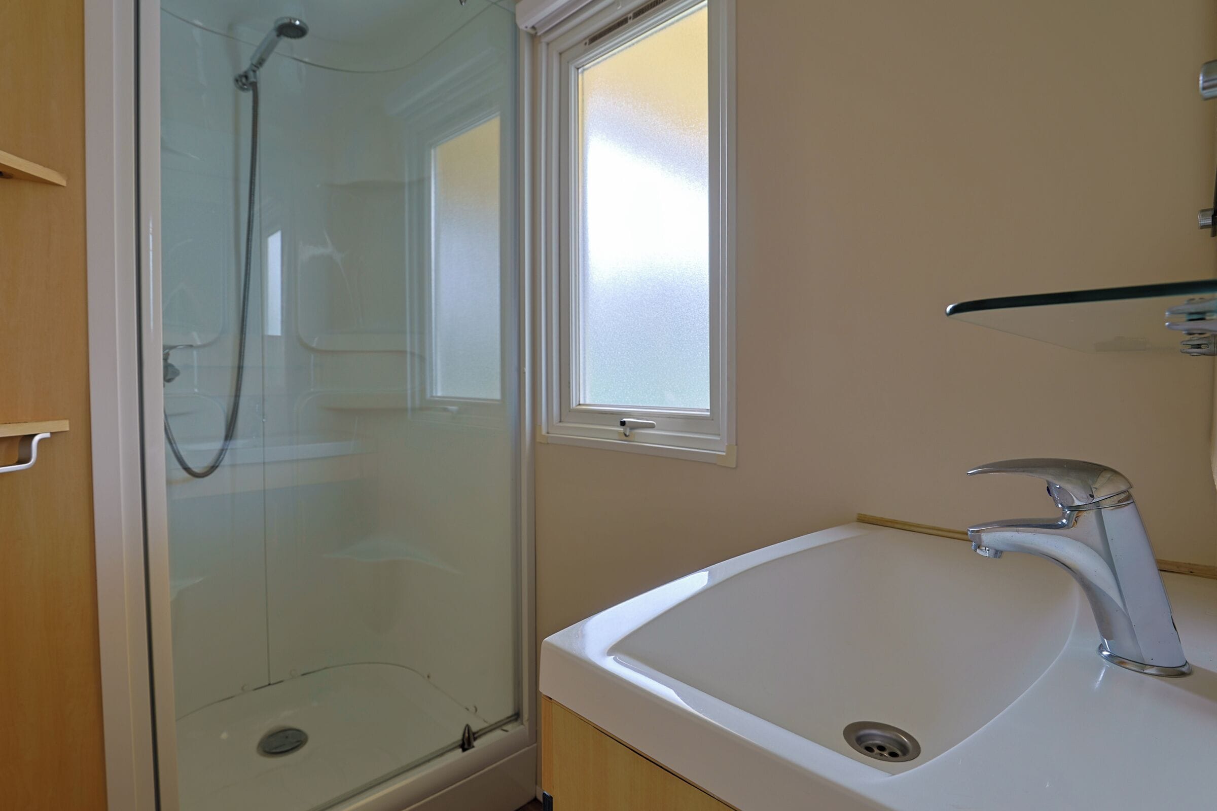 Een badkamer met een douche met glazen wanden, een wit wastafelmeubel met een moderne kraan, en een raam boven het wastafelmeubel.