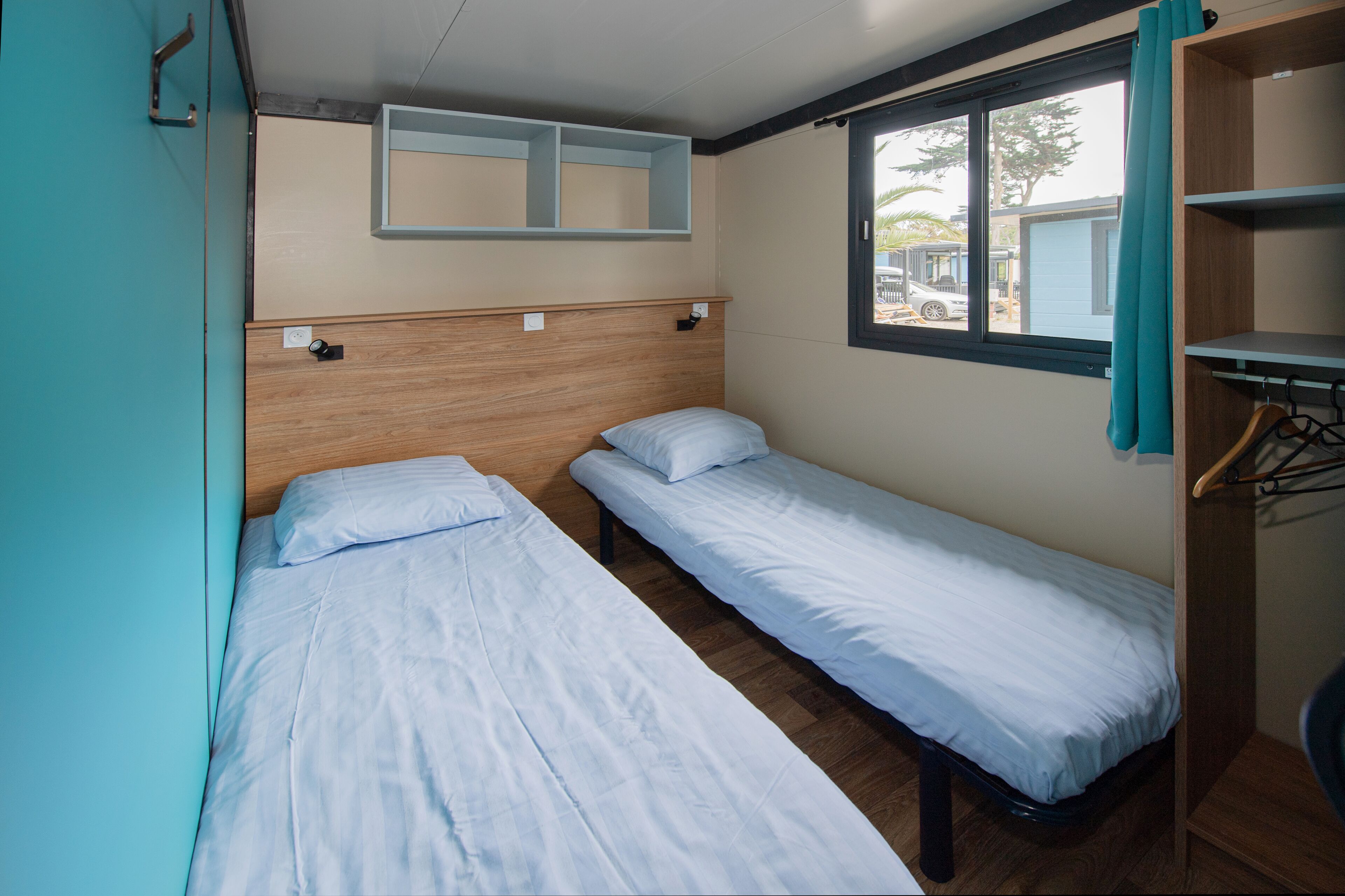 A small, compact room with two single beds, a wooden headboard, a window with a view of the outside, and a wardrobe with hanging space.