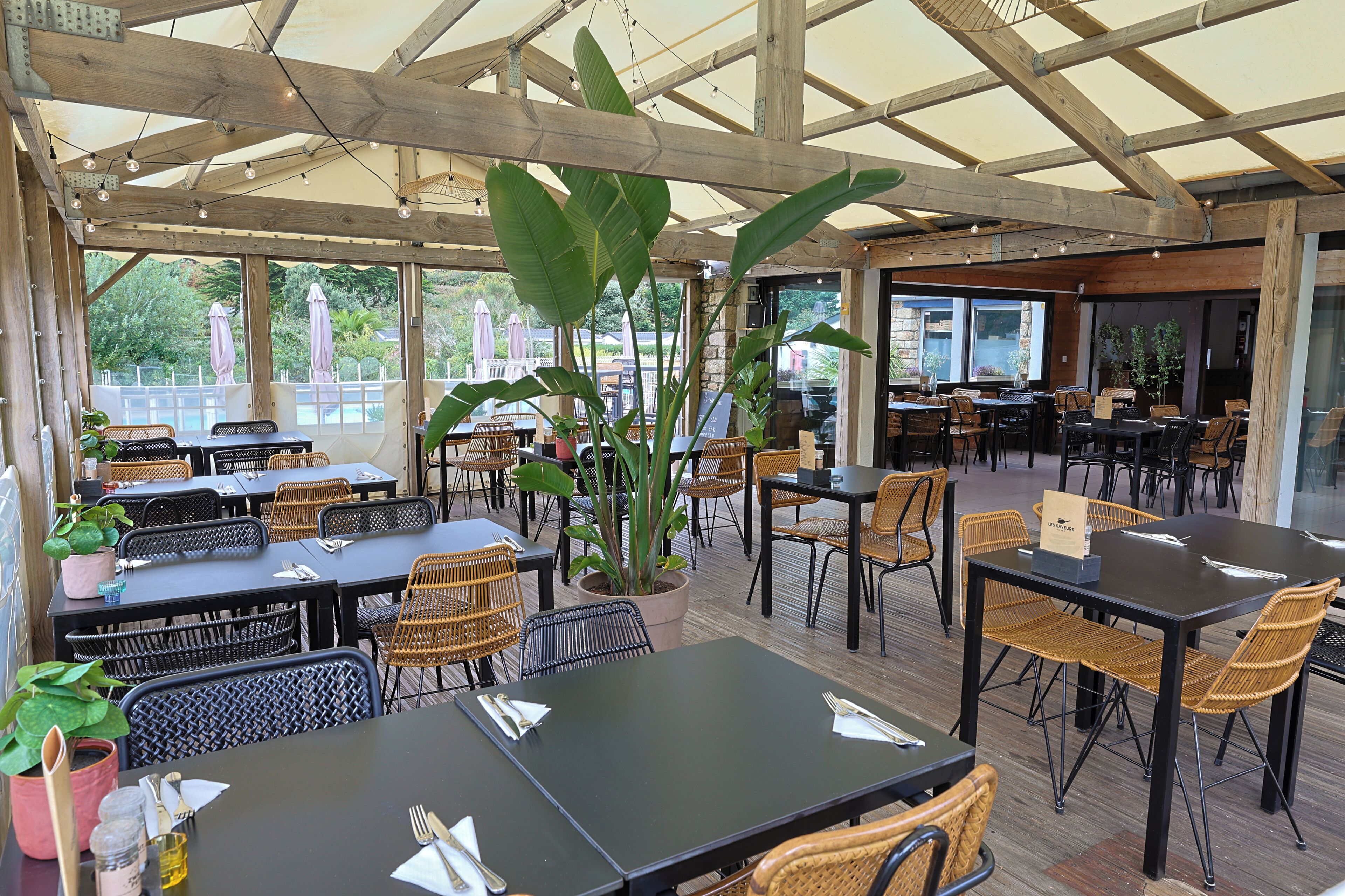 An outdoor cafe with wooden tables and chairs, surrounded by large plants and a view of the outdoors.