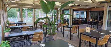 Un café al aire libre con mesas y sillas de madera, rodeado de plantas grandes y vistas al exterior.