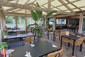 An outdoor cafe with wooden tables and chairs, surrounded by large plants and a view of the outdoors. - Camping RCN Port l'Épine (Trelevern)