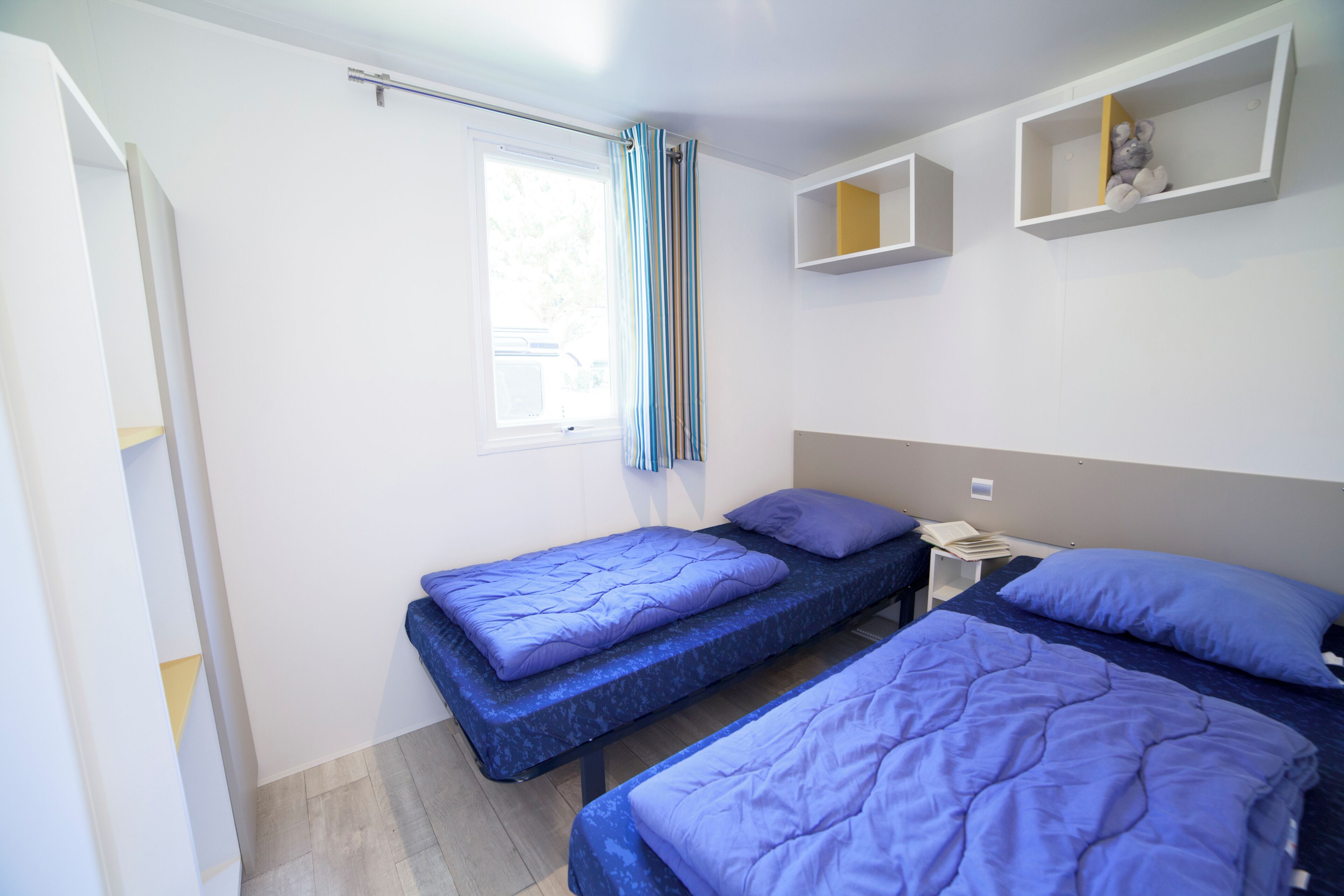 A bedroom with two beds, each with a blue mattress and matching pillows. There is a window with a curtain, a bedside table with a lamp, and a shelf with decorative items.