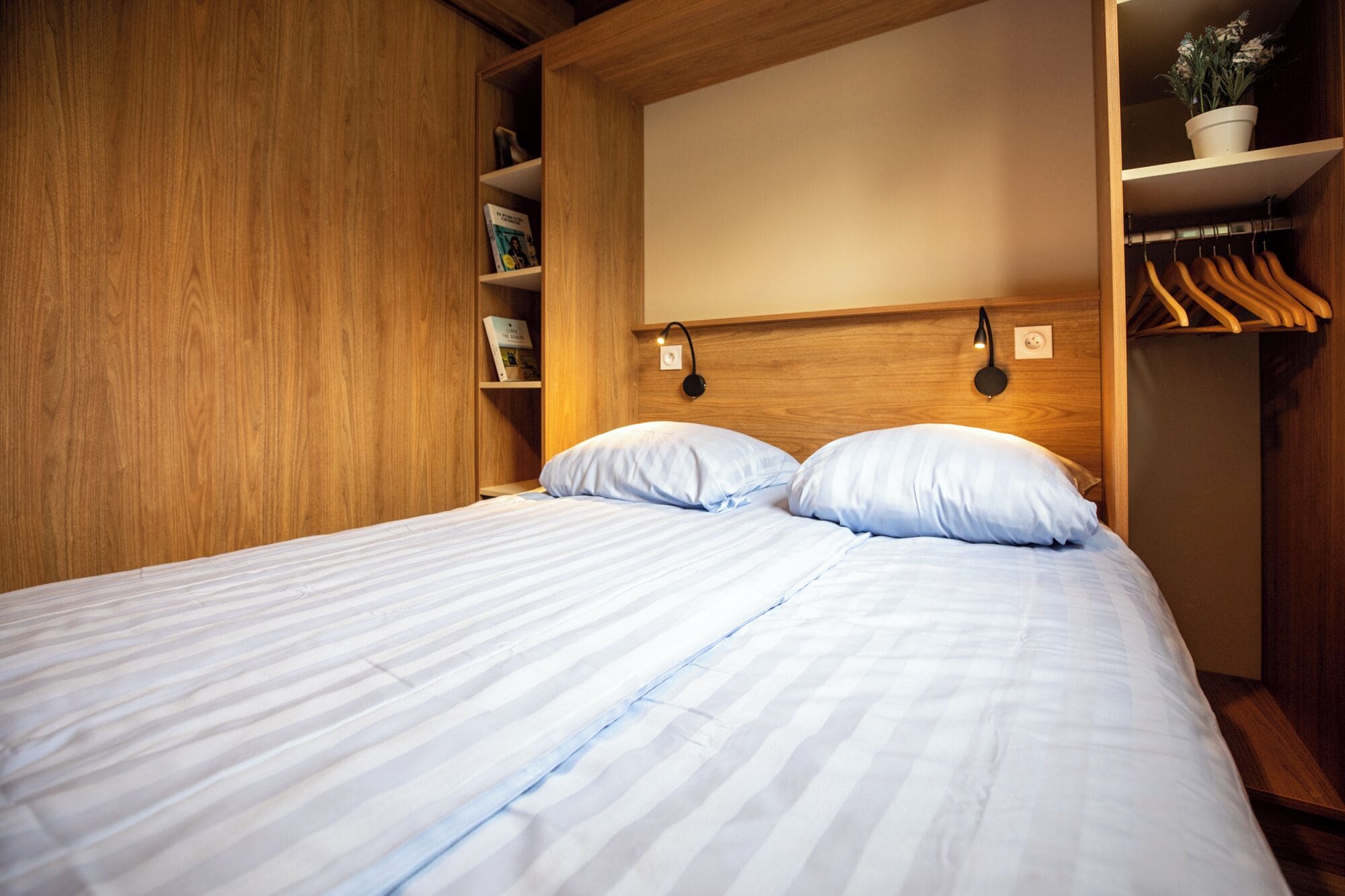 A neatly made bed with two pillows, a wooden wardrobe with a mirror, and a shelf with books and a potted plant.