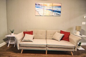 Un sofá color beige con patas de madera, dos cojines rojos y dos blancos, una mesita auxiliar con una planta, y un cuadro colgado en la pared que representa una escena de playa.