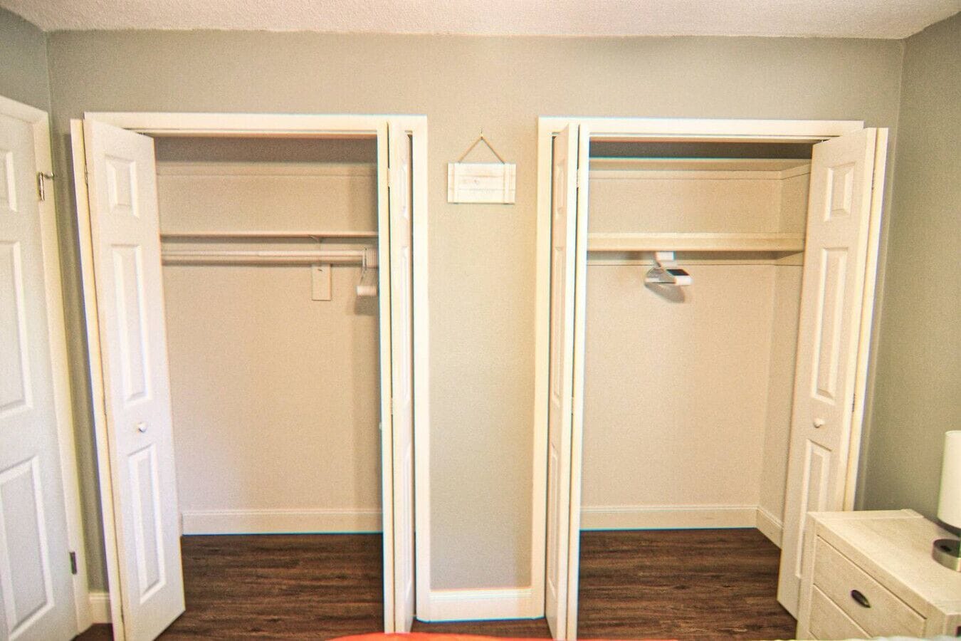 A room with two open closets, wooden flooring, and a small bedside table with a lamp.