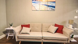 A beige sofa with wooden legs, two red and two white cushions, a small side table with a plant, and a wall-mounted painting of a beach scene. - The Cape 3 Gulf access (Cape Coral)