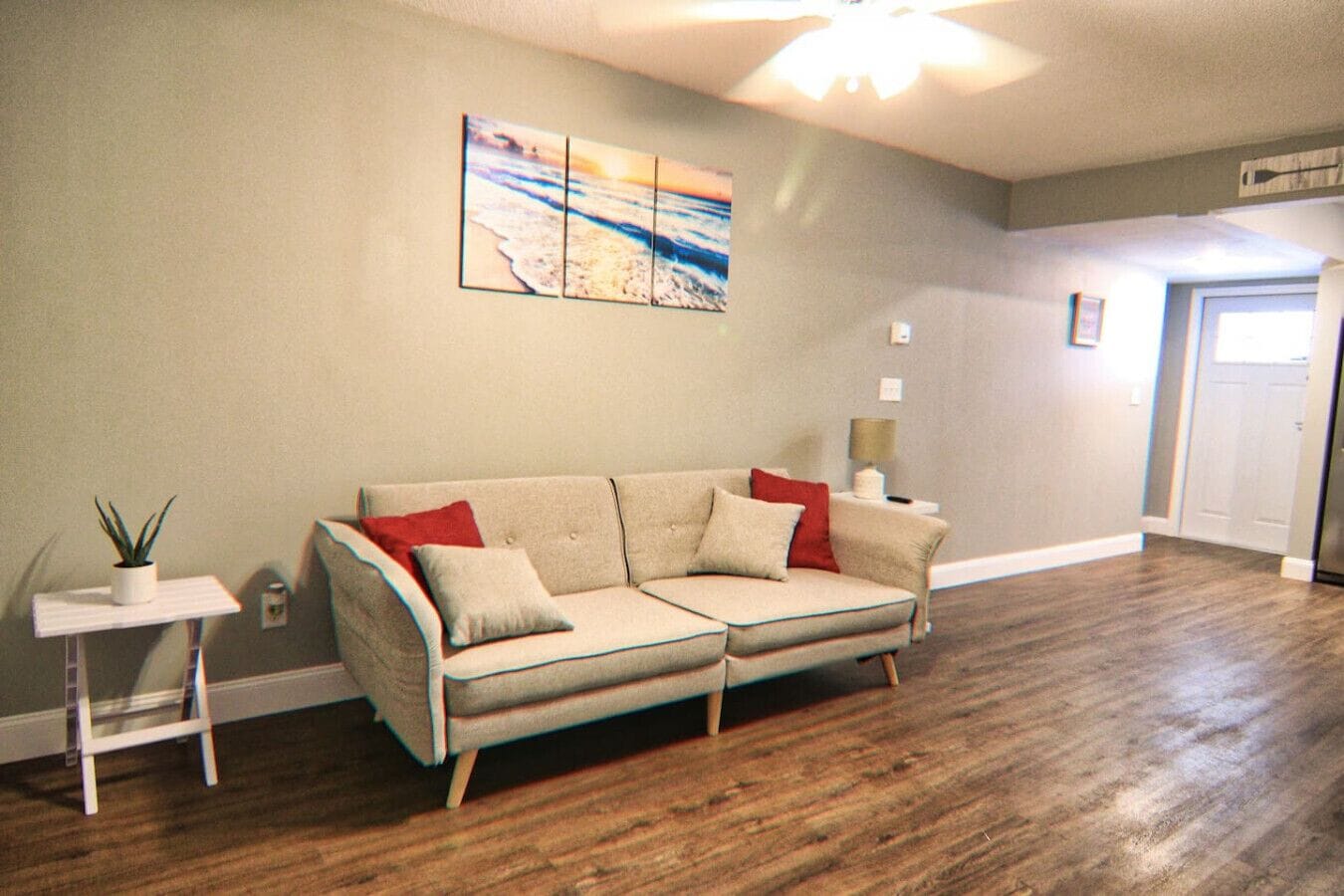 A living room with a beige sectional sofa, a small white side table with a plant, and a painting of a beach scene on the wall.