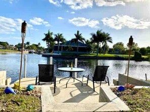 A waterfront patio with a table and chairs, overlooking a calm lake and a boat dock.
