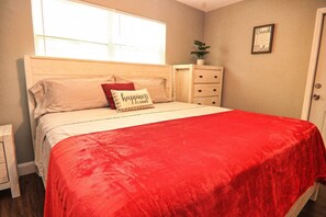 A bedroom with a bed, a red blanket, a pillow with 'happiness is home' text, a nightstand, a dresser, a window, and a door.