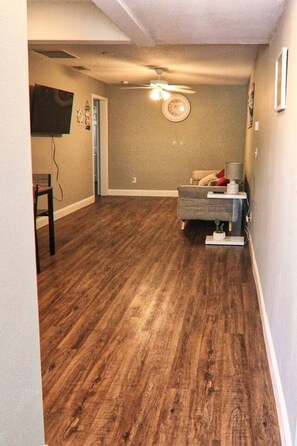 A room with hardwood flooring, a ceiling fan, a television mounted on the wall, and a small table with a lamp.