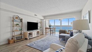 Una sala moderna con televisión, estanterías de madera, una zona de comedor y vista al mar.