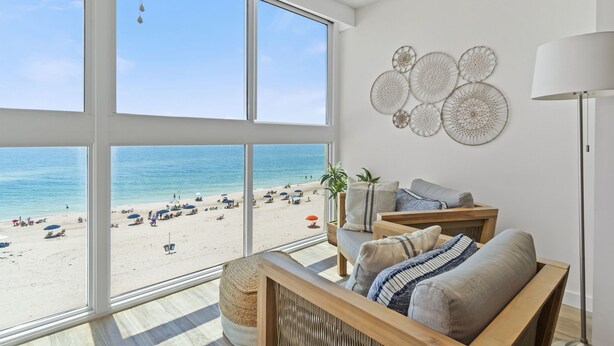 Una habitación con amplios ventanales que dan a una playa, un sofá de madera, una lámpara de pie y decoración en las paredes.
