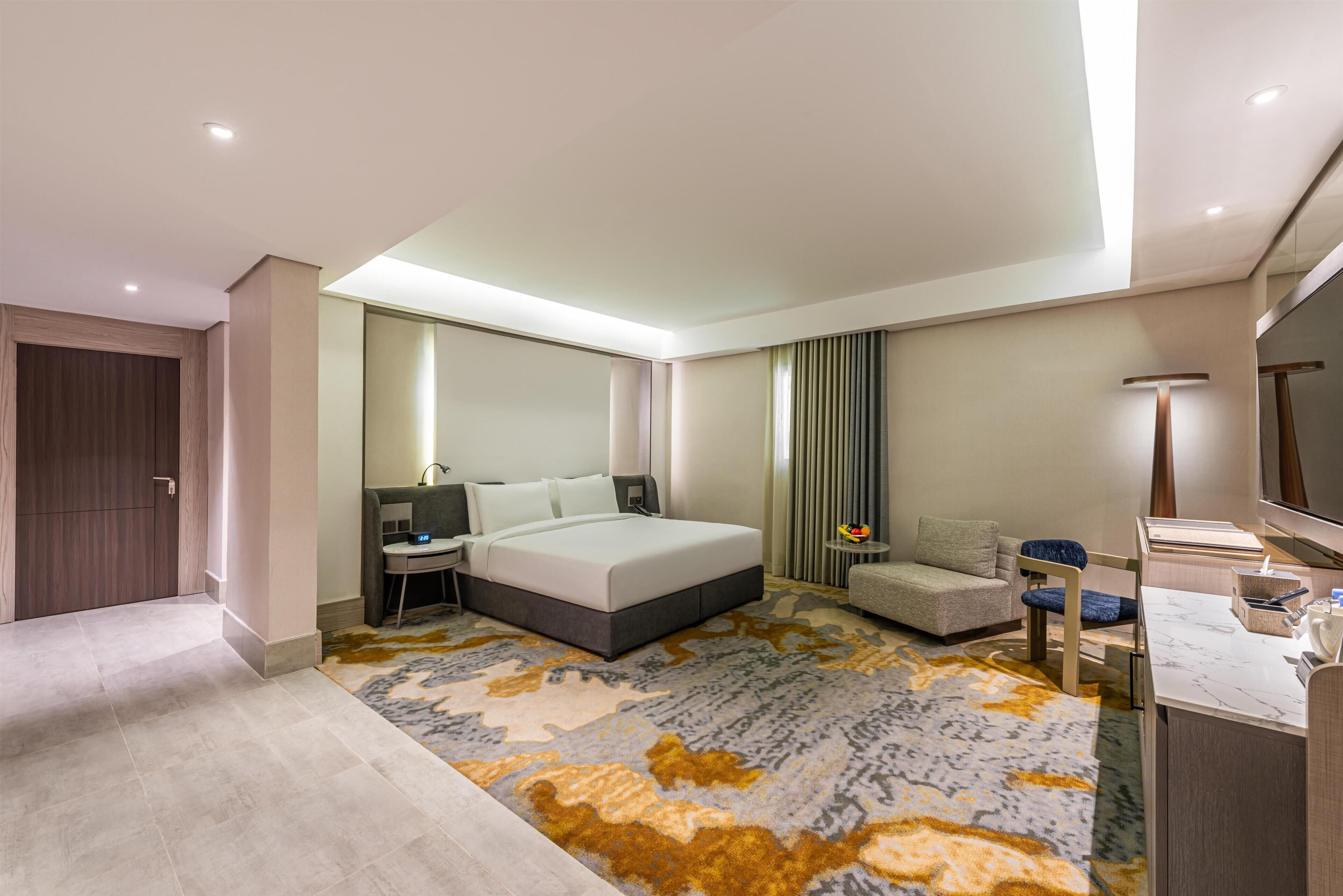 A modern hotel room with a large bed, a desk, a chair, a TV, and a bathroom area.