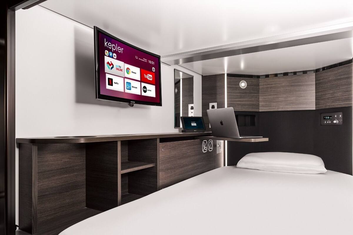 Una habitación de hotel moderna con cama, un escritorio con ordenador portátil, un televisor mostrando servicios de streaming y un sistema de sonido integrado.