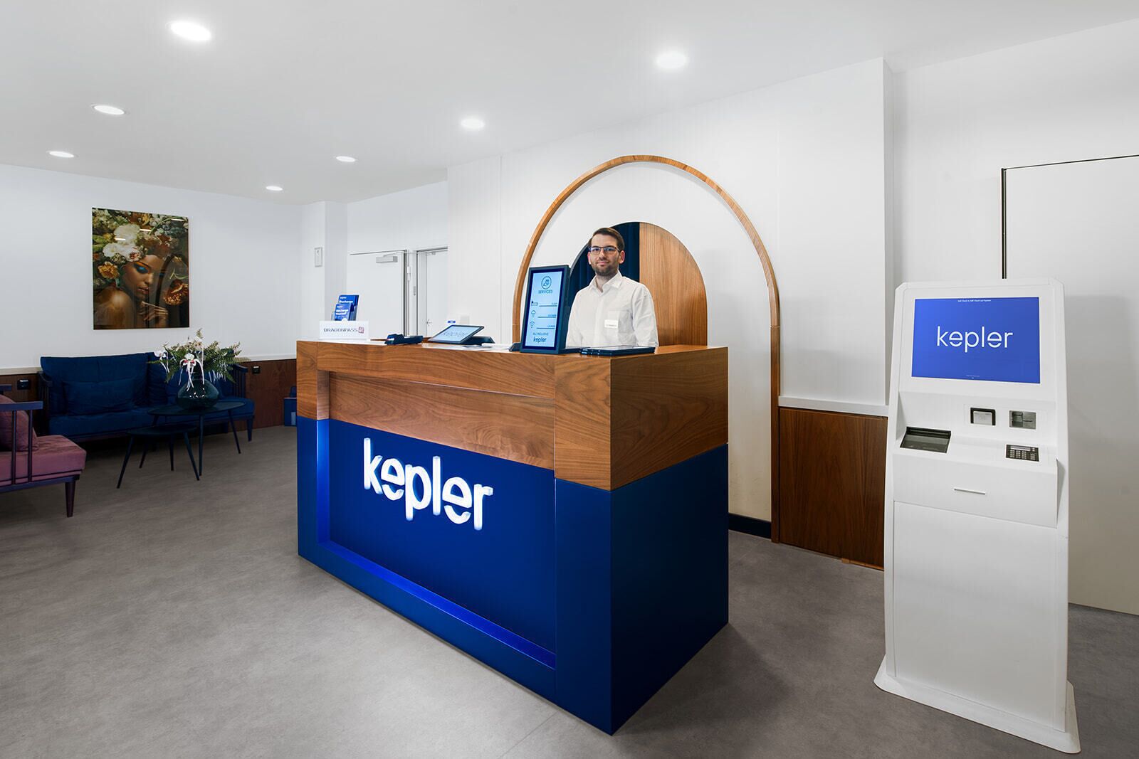 Vastaanottotiski, jossa on puinen kansi ja siniset sivut, digitaalinen infopiste sekä sininen kyltti, jossa on sana ”Kepler”.