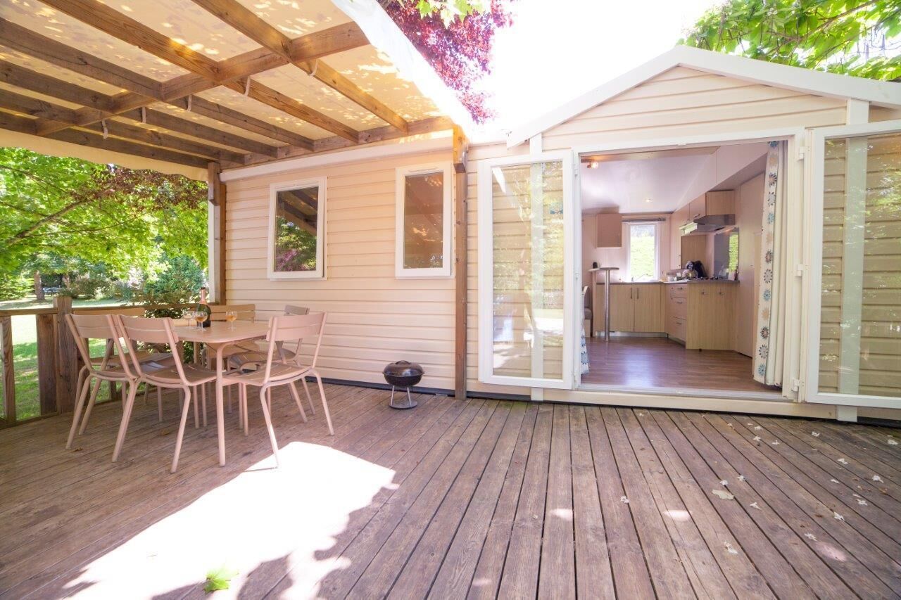 A wooden deck with a table and chairs, a covered patio area, and a view into a kitchen and living space.