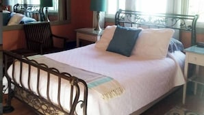 A bed with a metal frame, a bedside table with a lamp, and a chair in a room with a window and curtains. - Quaint Private Bungalow (Woodstock)