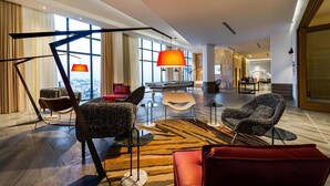 Un hall d’hôtel spacieux, doté de mobilier moderne, une grande fenêtre offrant une vue sur la ville et, au fond, un espace piscine.