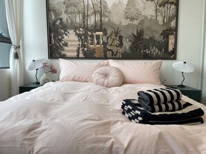 Ein ordentlich bezogenes Bett mit einer gestreiften Decke, zwei Nachttischlampen und ein großes gerahmtes Bild einer Gartenszene darüber.