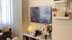 A cozy living room with a flat-screen TV mounted on the wall, a comfortable sofa, a coffee table with books and a lamp, and a bookshelf filled with books and decorative items. - City view of Melbourne GPO | Sunlit 2BR residence (Melbourne)