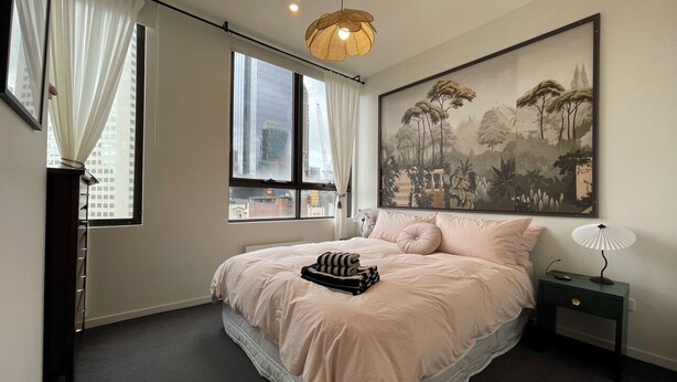 A bedroom with a large bed, a nightstand with a lamp, a window with curtains, and a painting on the wall. - City view of Melbourne GPO | Sunlit 2BR residence (Melbourne)