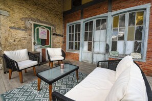 Una habitación con pared de ladrillo, un ventanal con marcos blancos y una puerta verde. Hay dos sillones blancos con cojines y un sofá de mimbre negro.